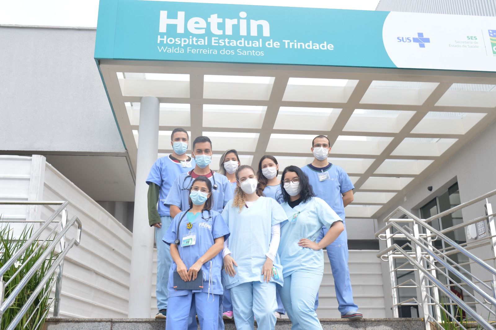Alunos de medicina, entre meninas e meninos estão à frente do hospital Hetrin nas escadas com roupas no tom azul claro, todos participam do Programa de Estágio Hetrin